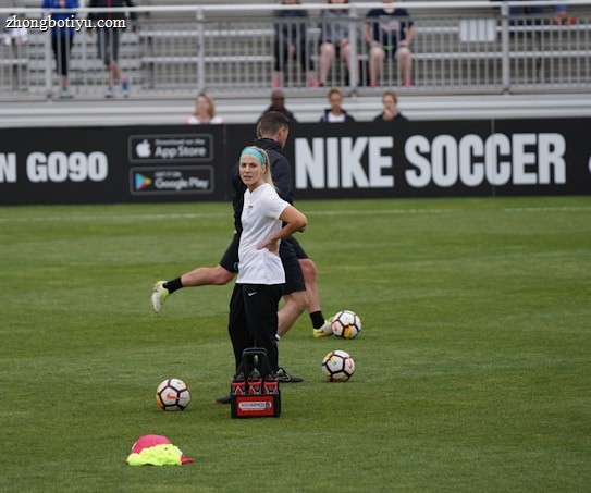 A person in sports attire stands on a soccer field, surrounded by several soccer balls and a carrier with bottles. The field is bordered by advertisements for Nike Soccer and app stores. Several people sit in bleachers in the background.