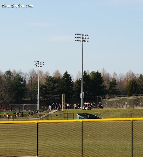 A sports field with a group of people engaging in athletic activities in the background. There is sports equipment like a soccer goal and a scoreboard visible. A chain-link fence runs across the foreground, and a statue or sculpture resembling an eagle is also seen. Trees and tall floodlights are positioned behind the field.