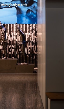 A dimly lit display features statues of athletes standing on a podium. They are positioned in front of a backdrop with the words 'LEVELING THE PLAYING FIELD' in bold letters. On the wall to the right, a softly lit quote is partially visible. The overall ambiance suggests a museum or exhibition setting.