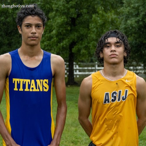Two young athletes wearing sleeveless sports tops stand on a grassy field. The athlete on the left is wearing a blue top with the word 'TITANS' while the one on the right wears a yellow top with 'ASIJ'. The background features green trees and a white fence, with a bench visible on the right.